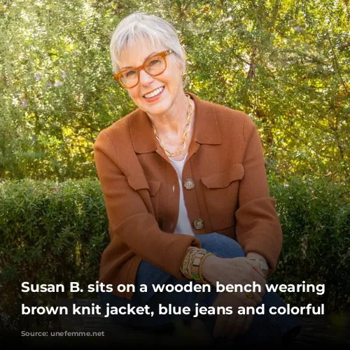 Susan B. sits on a wooden bench wearing a brown knit jacket, blue jeans and colorful bracelets.