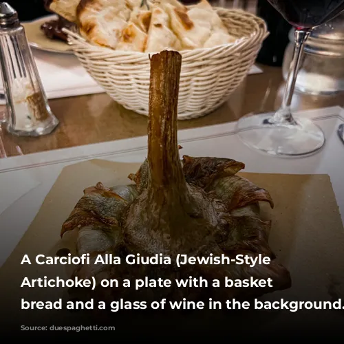 A Carciofi Alla Giudia (Jewish-Style Fried Artichoke) on a plate with a basket of bread and a glass of wine in the background.