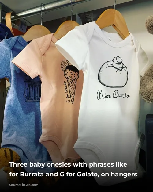 Three baby onesies with phrases like B for Burrata and G for Gelato, on hangers 