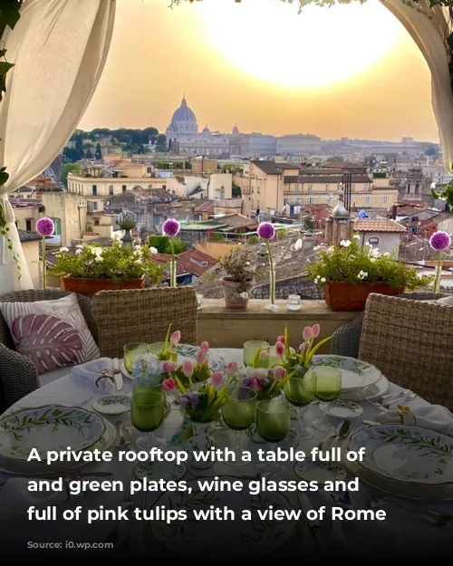 A private rooftop with a table full of white and green plates, wine glasses and vases full of pink tulips with a view of Rome