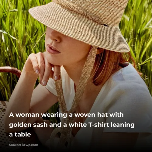 A woman wearing a woven hat with a golden sash and a white T-shirt leaning on a table 