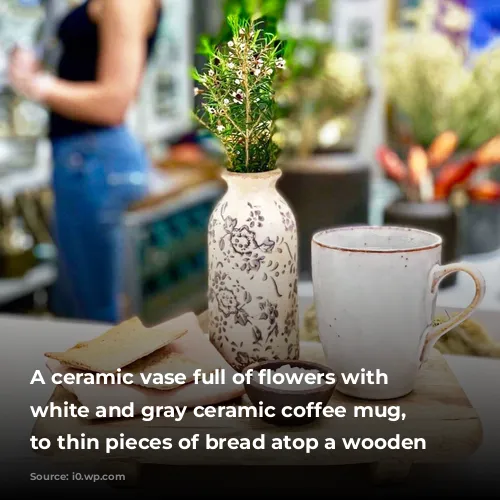 A ceramic vase full of flowers with a white and gray ceramic coffee mug, next to thin pieces of bread atop a wooden board